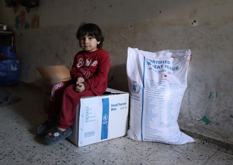 A young child sitting in an emtpy room on a food parcel next to a bag of wheat flour provided by WFP. © WFP/Ali Jadallah