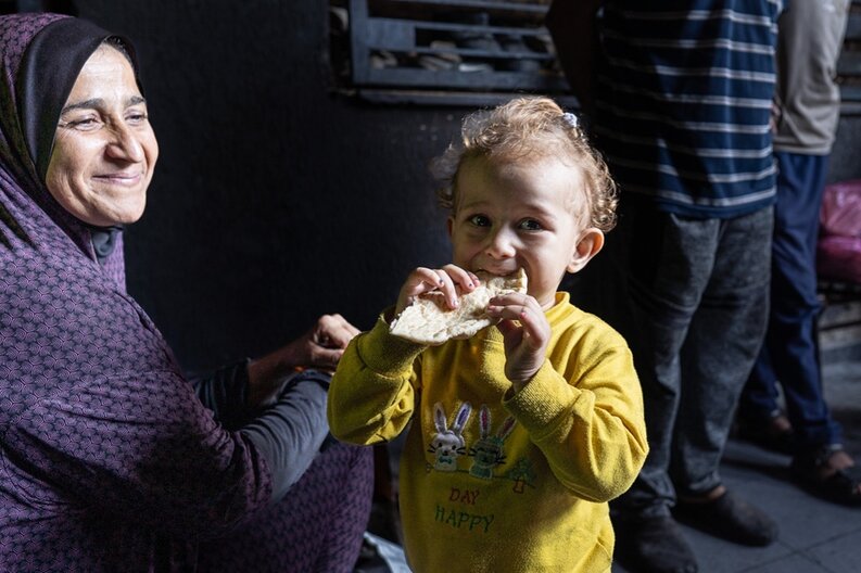 A young child eating bread and standing next to its mother.