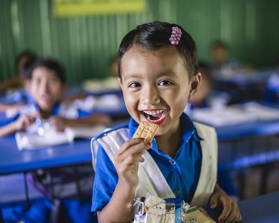 A girl sits in a classroom holding a biscuit with a huge smile on her face