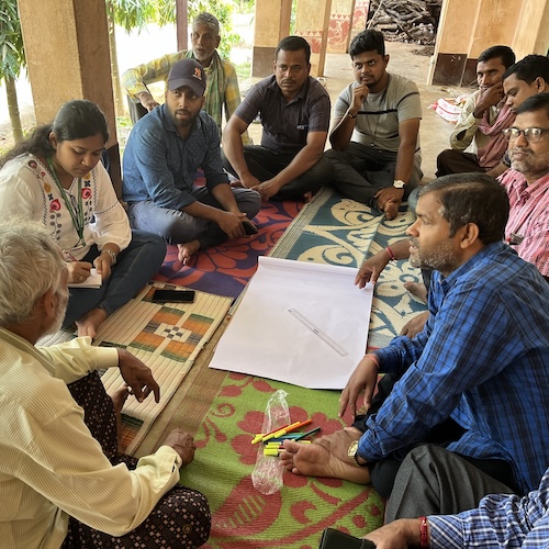 Group seated on mats discussing around a large sheet of paper with markers and notes in an open-air setting.
