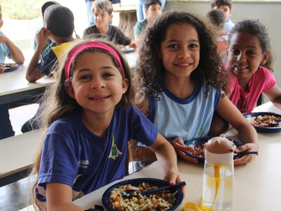 Three children sat at a table and smiling, each with a plate of food. Behind them are children eating at another desk.