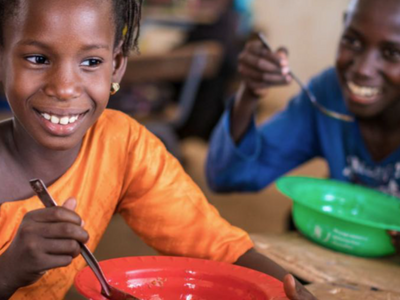 Photo: WFP Child eating a meal from a red bowl in a classroom feeding programme, while another child eats in the background, highlighting school meals support.