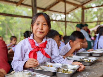 Students sit together at long wooden tables in an open-air school dining area, eating a meal served in metal trays. A student in the foreground holds a spoon while other students eat and talk in the background, illustrating a school meals programme.