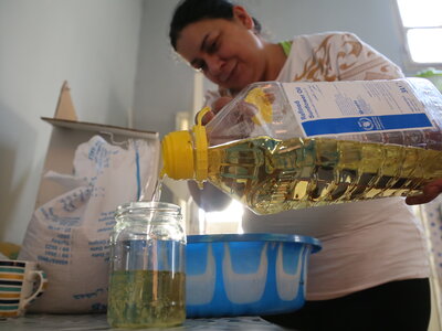 A person pouring cooking oil from a large, WFP-marked plastic bottle into a glass jar in a kitchen, with a striped mug and bag on the table.