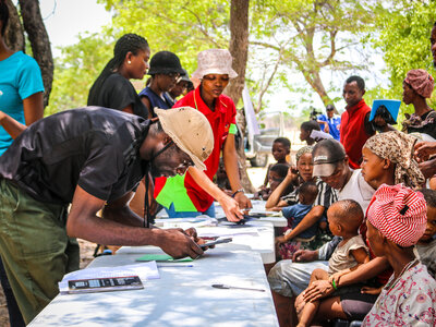 People gather around a table outdoors as staff register households using mobile devices and paper forms, while caregivers sit with children nearby under trees.
