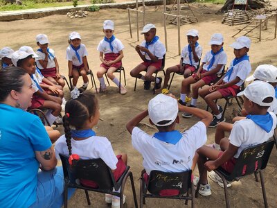 Photo: WFP Children wearing white caps and blue scarves sit in a circle outdoors with WFP staff, participating in an interactive group activity.