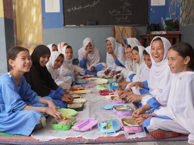 Schoolchildren sit together on floor mats inside a classroom, eating a shared meal from lunch containers. A blackboard and classroom furnishings are visible in the background.
