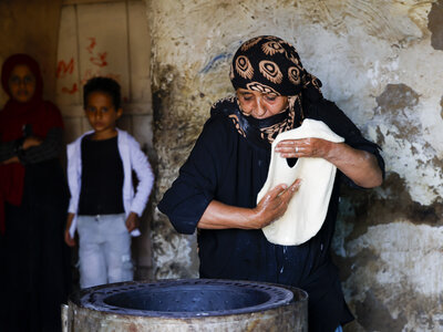 A person in a patterned headscarf stretches dough over a cooking surface, with two others standing in the background against a textured wall.