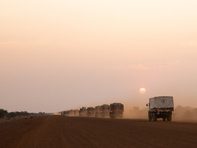 A convoy of trucks drives on a dusty road at sunset, the sun low on the horizon casting long shadows under a clear sky.