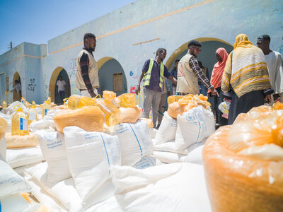 People gather around food supplies labeled 'World Food Programme' with cooking oil containers, outside a building with arched doorways.