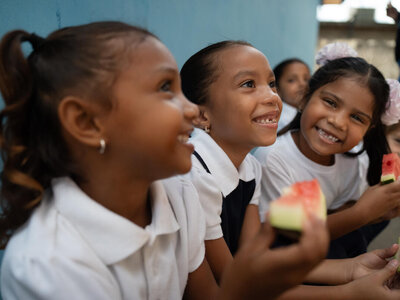 Children in school uniforms sit against a blue wall, holding slices of watermelon in their hands.