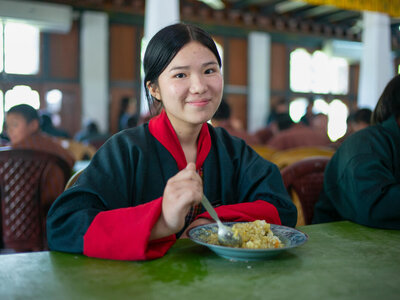 Photo: WFP/Rein Skullerud Student in traditional dress sits at a dining table with a plate of food, holding a spoon in a busy school cafeteria with other diners behind.