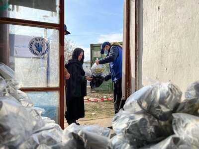 A man in a blue WFP vest and face mask distributes food to a woman in a black headscarf outside a distribution centre, with bags of supplies in the foreground and a WFP logo visible on the glass door. 