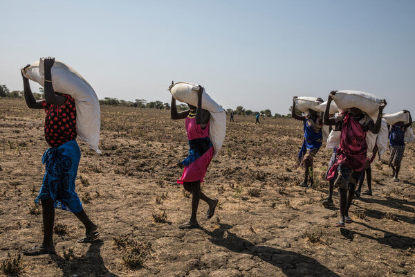 women walk on barren ground carrying bags of food over their head and shoulders