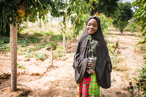 A girl stands in a school garden holding a small plant with a big smile