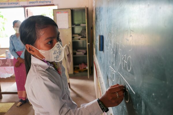A girl wearing a face mask writes on a blackboard while her teacher watches