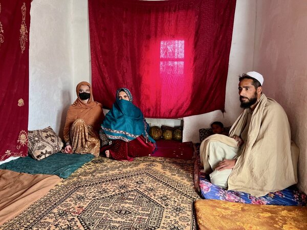 A man and two women in traditional clothes sit in a room decorated with pillows, carpets and mattresses. 