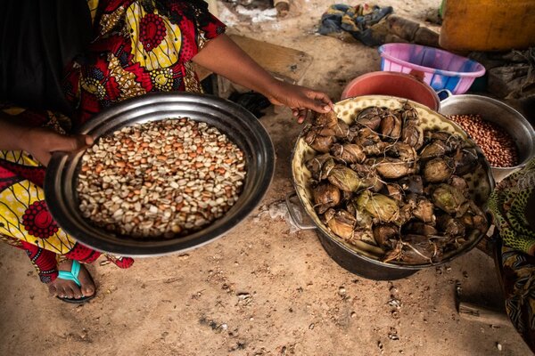 A woman displays a bowl of mixed grains beside a large container filled with wrapped food items on the ground in an outdoor cooking area.