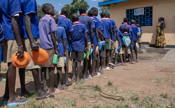 Children in blue uniforms and carrying plastic containers line up for food in a dusty yard. Photo: WFP/Alessandro Abbonizio