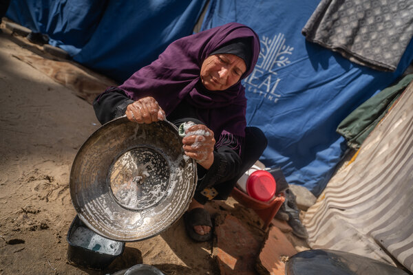 A woman kneels on the ground in a makeshift shelter, scrubbing a metal pot with soapy water beside basic household items.