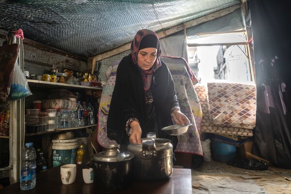 Woman in a modest shelter kitchen preparing food beside metal pots, with shelves of supplies and folded bedding in the background.