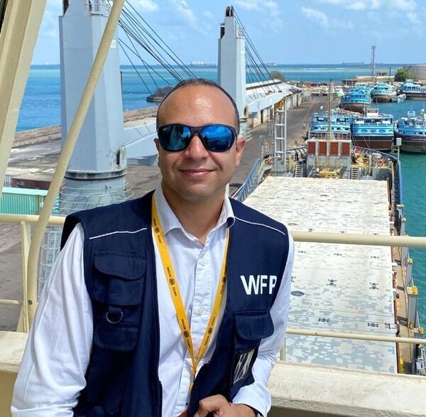 WFP staff member wearing a blue vest stands on a ship deck overlooking a busy port, with cranes, moored boats and turquoise sea under clear skies. 