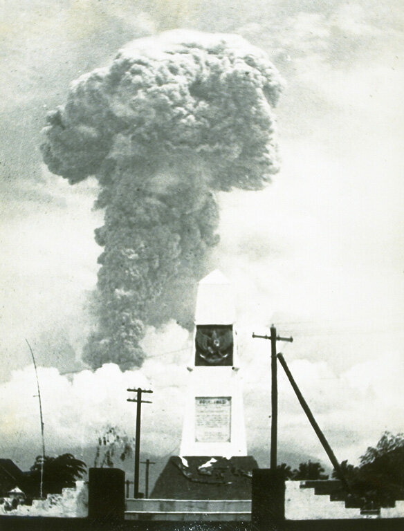 Large volcanic ash plume rising into the sky behind a monument and utility poles, with buildings at ground level in the foreground.