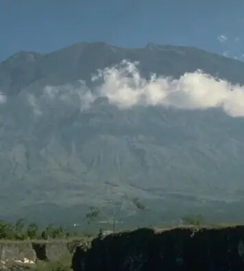 Mountain volcano rising above a low green landscape, with a thin band of clouds drifting across the mid-slope under a clear blue sky.