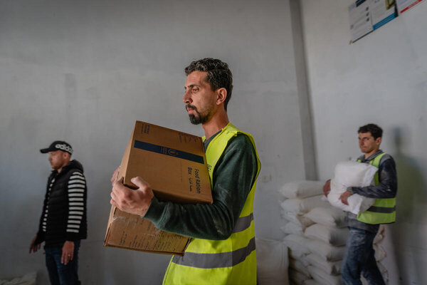 Aid workers in high-visibility vests carry food boxes and sacks inside a warehouse, with stacked supplies and others moving goods nearby.
