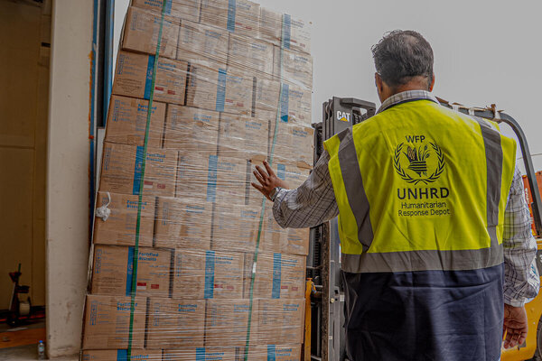 Forklift operator lifts a tall pallet of WFP fortified biscuits wrapped in plastic, stacking boxes inside a warehouse with organised food aid supplies.