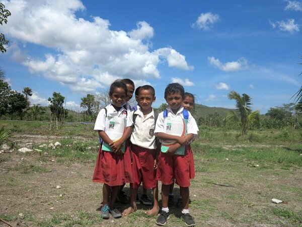 Five schoolchildren stand outdoors in a grassy rural area, wearing uniforms and holding notebooks, with trees and hills under a bright blue sky.
