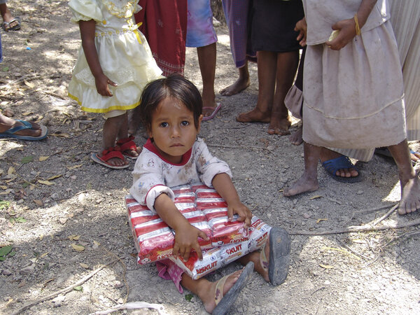 Child sits on ground holding a bundle of packaged biscuits while other children stand nearby on a dry, dusty clearing.