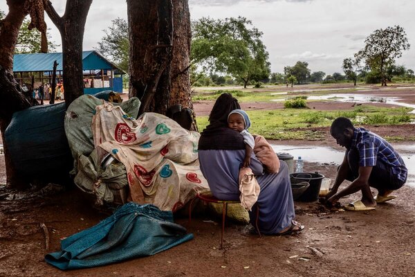A man in a plaid shirt prepares food outside, while a woman cradling a small child sits on the muddy ground behind him. Photo: WFP/Gabriela Vivaqua