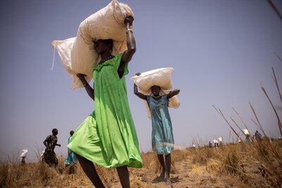 A woman in a green dress carries a white bag of food over her head in a field in South Sudan