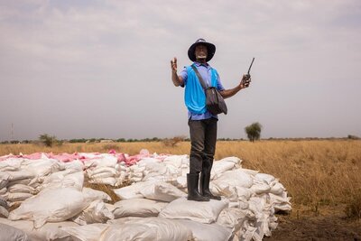 A man in a WFP vest stands on top of neatly stacked bags of food in a field in South Sudan, holding a radio