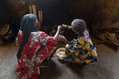 Person and child prepare food over a small fire inside a dimly lit home, stirring a cooking pot beside a bowl of flour.
