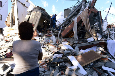A person stands before the rubble of a collapsed building as rescue workers search through debris and twisted concrete under a blue sky.