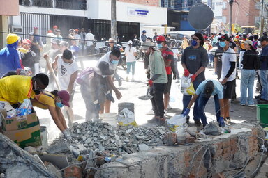 People clear rubble from a damaged urban area in Cali, Colombia, working together with shovels and buckets amid dust and debris. 