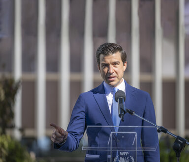 Person speaking at a WFP podium outdoors during an official event, wearing a blue suit and standing behind a microphone with a modern building in the background.