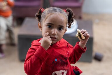 A little girl in a red shirt in Gaza eating a piece of bread.