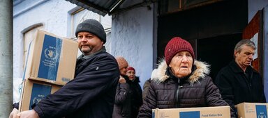 People carry WFP food boxes outside a warehouse, transporting aid supplies as part of a distribution effort in a cold weather setting.