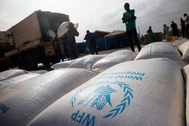 WFP food bags stacked as workers unload supplies from a truck at a distribution site.