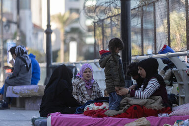 Families rest on blankets along a street, with adults and children sitting near personal belongings beside barriers and fencing during displacement..
