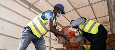 WFP workers wearing high-visibility vests unload boxes of humanitarian supplies from a truck, transferring cartons by hand during logistics operations.