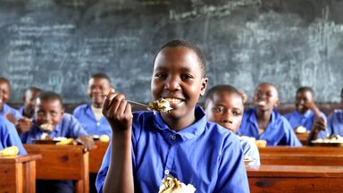 WFP/Aristide Gatera A child at a school desk with a fork-full of food