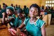 A smiling young schoolgirl wearing a green uniform sits in a classroom with other students. Photo: WFP/Michael Duff