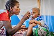 A woman in a grey and red T-shirt spoons food into the mouth of her baby, who has been hospitalized with malnutrition. Photo: WFP/Charly Kasareka