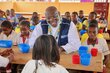 A man wearing a WFP jacket and African beads sits in a primary school classroom in Cameroon chatting with kids. Photo: WFP/Jordan Onana