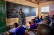 A woman in a dark patterned dress reads out sentences on a blackboard to a classroom of young students. Photo: WFP/Alessandro Abbonizio 
