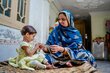 A mother in a long blue headscarf and robe hands a smiling little girl in a yellow dress a package of specialized nutritional food. Photo: WFP/Sahil Zia